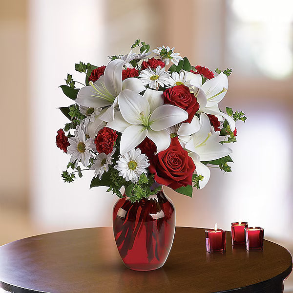 Bouquet of red and white flowers in a red vase on a wooden table with candles.
