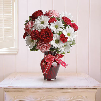 Bouquet of red, white, and pink flowers in a red vase with a bow on a white surface.