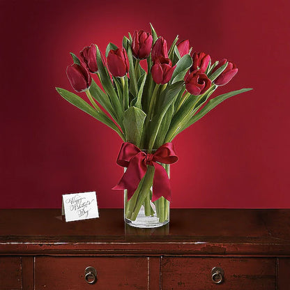 Bouquet of red tulips in a clear vase with a red bow on a wooden surface against a red background.